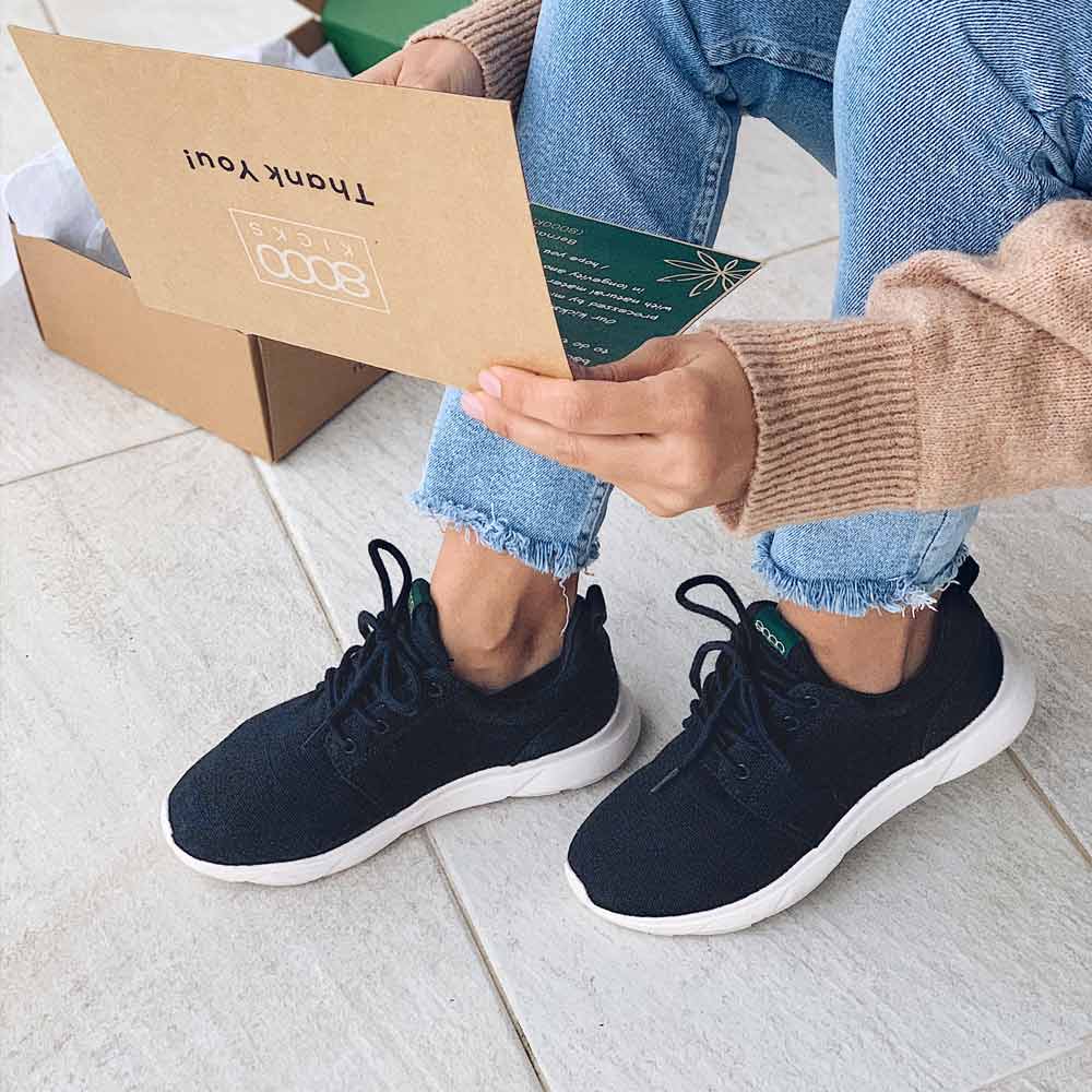 Person sitting indoors wearing the 8000 Kicks Low Top Explorer V2 Black and White sneakers while reading a thank you note. The sustainable sneakers are paired with light blue jeans and a beige sweater.