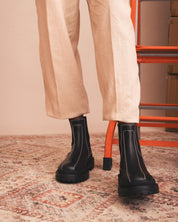 Person wearing black boots and beige pants on a patterned rug with an orange chair in the background.