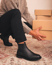 Person wearing black boots and holding cardboard boxes on a patterned rug.