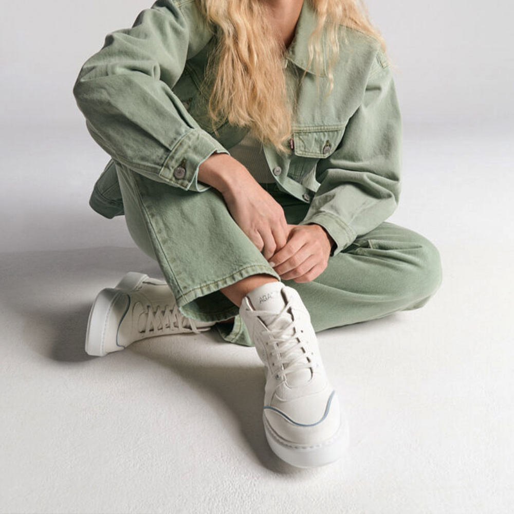 Model wearing AGAZI EMI High Top sneakers in white and blue, seated pose. Sustainability shoes. Environment friendly shoes.