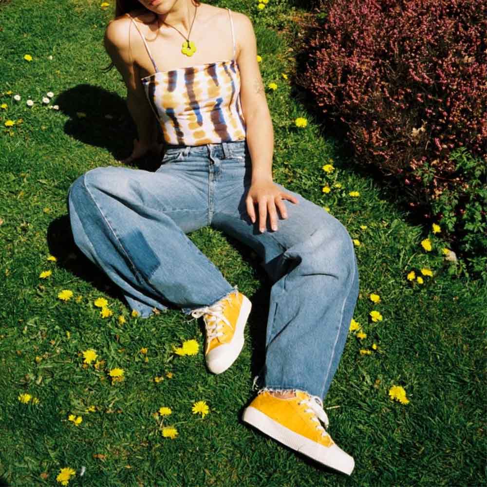 Woman sitting on grass, wearing Bamburista yellow sneakers paired with jeans and a tie-dye top, highlighting the sustainable shoe in a natural setting.