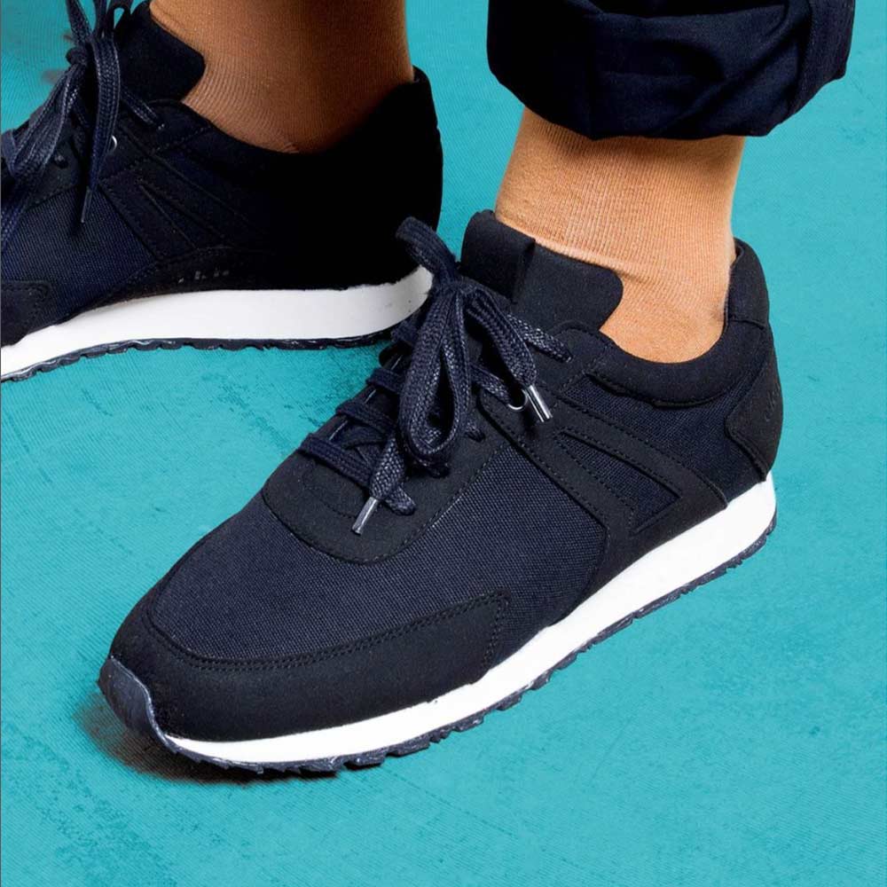 Model wearing the EKN Low Seed Runner sustainable sneaker in black standing on a turquoise floor and wearing orange socks.