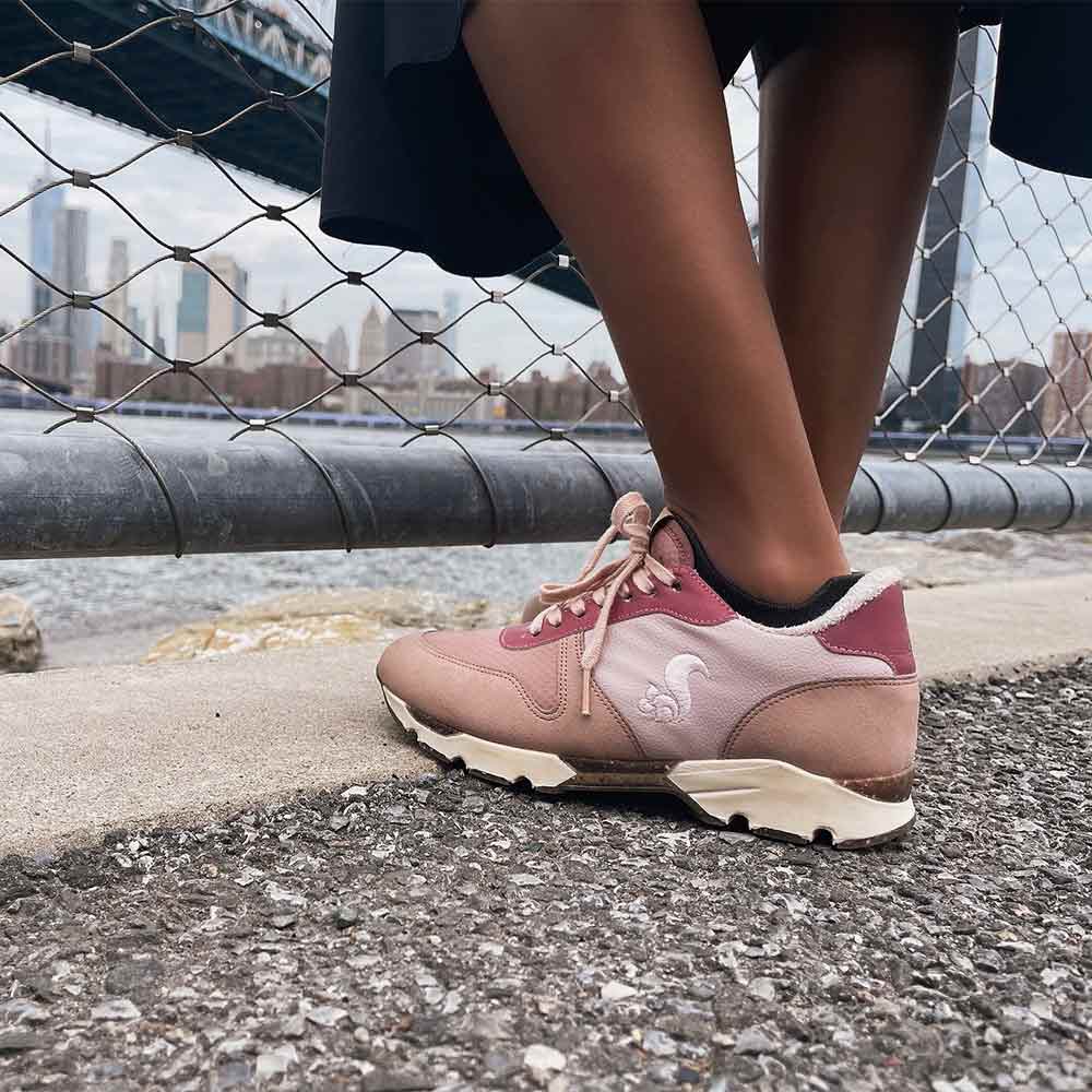 A person wearing Thies Sustanable Vegan shoes, Eco Squirrel Runner in rose color, standing on a paved surface by a waterfront, with a cityscape and bridge in the background.