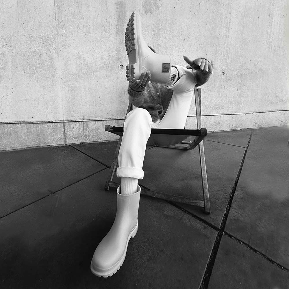 Black and white image of a person wearing Thies Bio Rain Boots, sitting on a chair and showcasing the vegan boots.