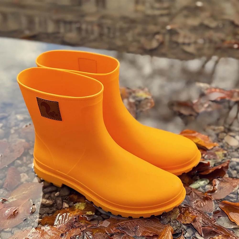 A pair of Thies Bio Rain Boots in bright orange, designed for women, showcased on a wet surface with fallen leaves, highlighting their vegan and waterproof features.