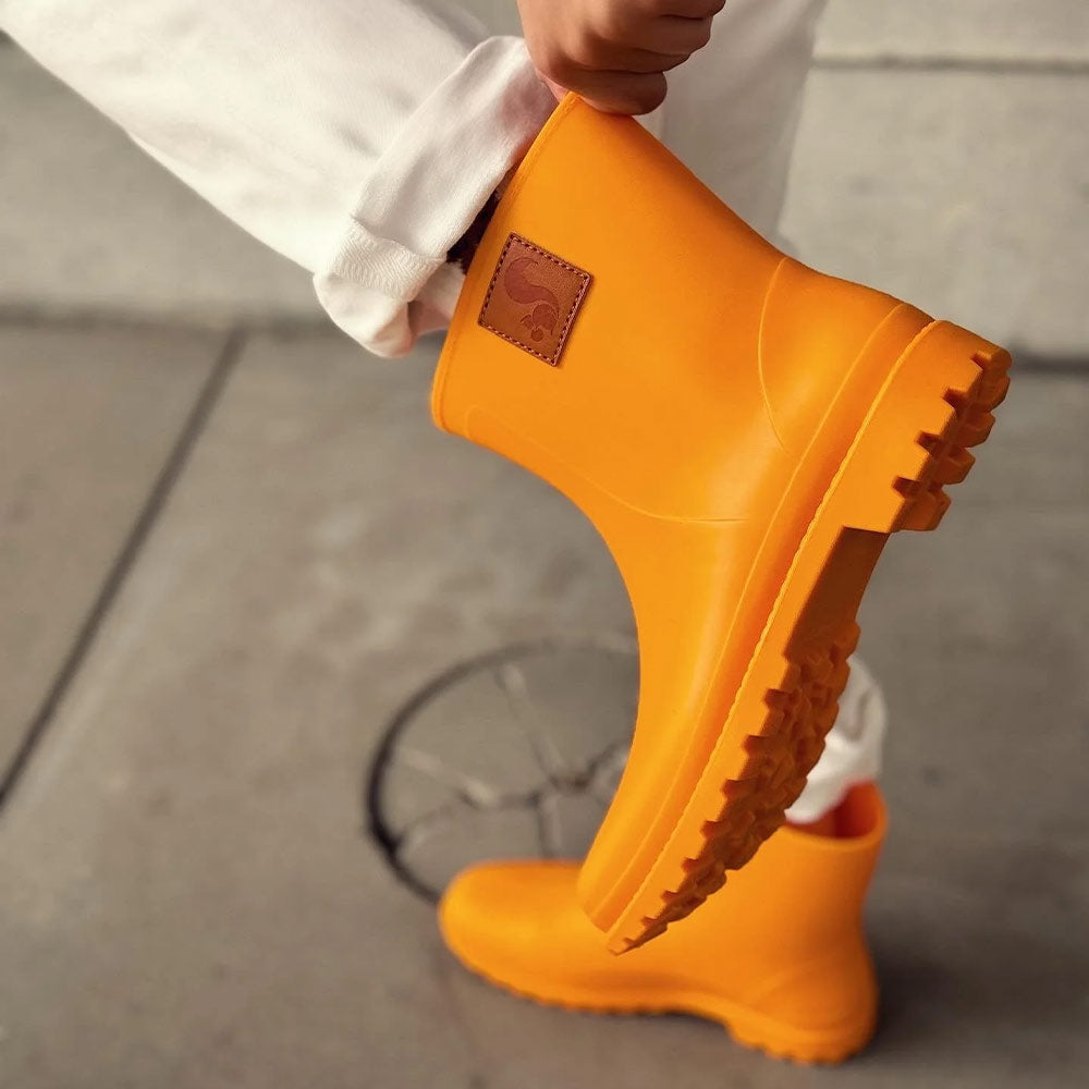 A close-up of a woman adjusting her Thies Bio Rain Boots in bright orange, emphasizing their vegan-friendly materials and sturdy construction.