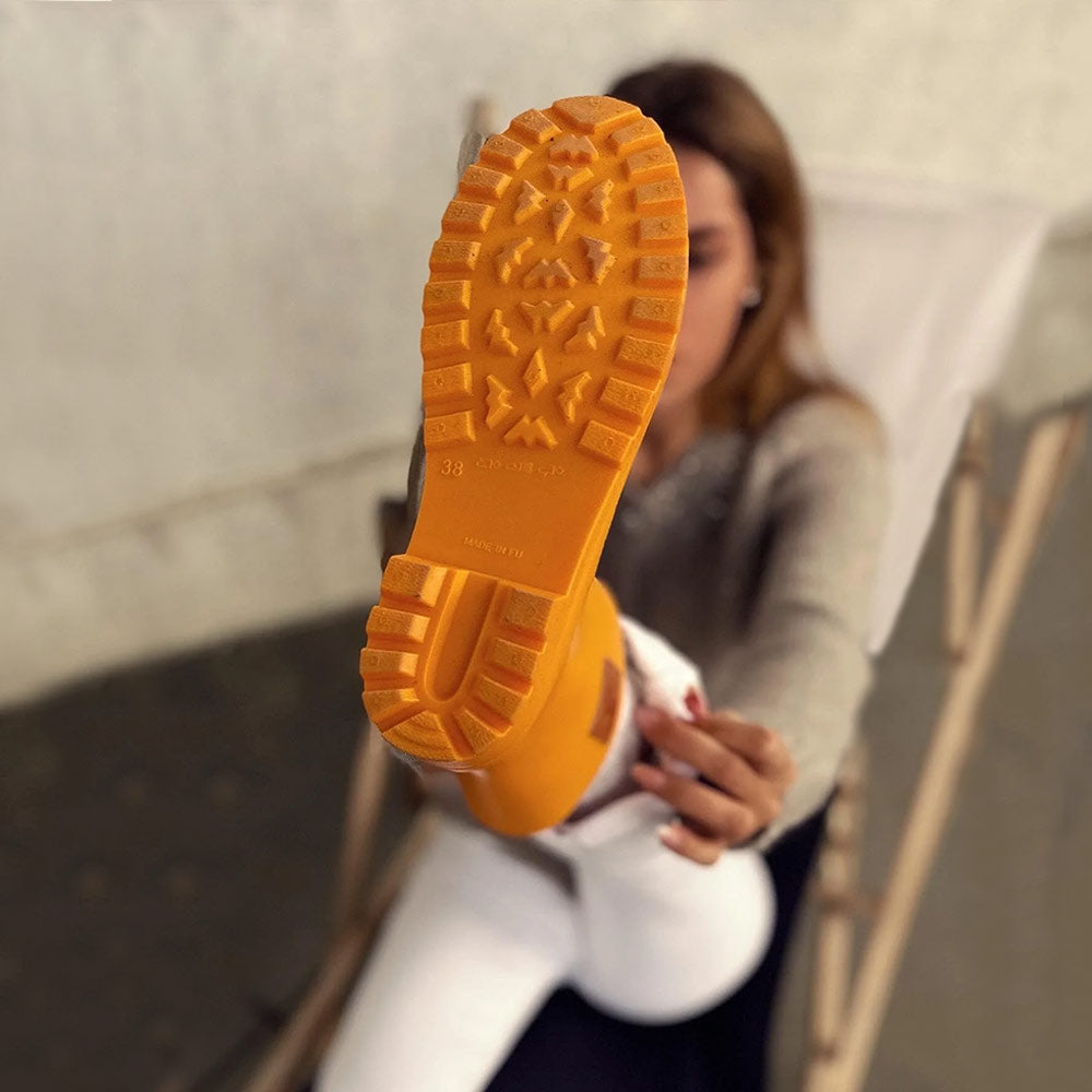 The sole view of a Thies Bio Rain Boot in bright orange, worn by a woman sitting and showing the detailed tread pattern, illustrating the boot's vegan and durable design.